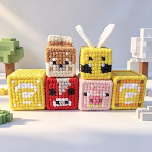 May include: A collection of six crocheted cube-shaped plush toys featuring pixelated designs. The set includes two yellow question mark blocks, a brown fox, a red block with a face, a pink pig, and a yellow bee with white yarn wings.