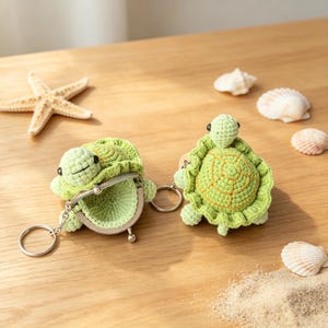 Op de afbeelding: Twee lichtgroene gehaakte schildpad-muntbeursjes met metalen knipsluitingen en zilverkleurige sleutelringen. Elke schildpad heeft een textuurrijk schild met oranje stiksels, kleine zwarte oogjes en een zachte, ronde lichaamsvorm.
