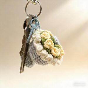 K&ouml;nnte beinhalten: Ein handgefertigter H&auml;kel-Schl&uuml;sselanh&auml;nger mit einem Strau&szlig; gelber und gr&uuml;ner Blumen. Das Bouquet ist von einem wei&szlig;en und grauen H&auml;keldesign umgeben. Der Schl&uuml;sselanh&auml;nger ist an einem silbernen Schl&uuml;sselring mit mehreren Schl&uuml;sseln befestigt.
