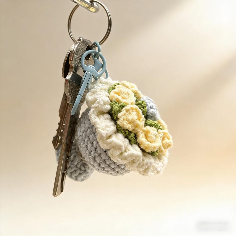 K&ouml;nnte beinhalten: Ein handgefertigter H&auml;kel-Schl&uuml;sselanh&auml;nger mit einem Strau&szlig; gelber und gr&uuml;ner Blumen. Das Bouquet ist von einem wei&szlig;en und grauen H&auml;keldesign umgeben. Der Schl&uuml;sselanh&auml;nger ist an einem silbernen Schl&uuml;sselring mit mehreren Schl&uuml;sseln befestigt.