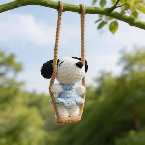Könnte beinhalten: Eine gehäkelte Snoopy-Puppe in einem hellblauen Kleid, auf einer Schaukel aus braunem Seil. Die Puppe hat schwarze Ohren und eine schwarze Nase. Die Schaukel hängt an einem Ast. Der Hintergrund ist verschwommen grün.