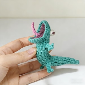 Può includere: Una piccola statuetta di lucertola all'uncinetto color verde acqua, caratterizzata da una bocca spalancata con interno viola e una lingua rosa sporgente. La creatura ha un occhio bianco visibile con pupilla scura, arti piccoli e una lunga coda.