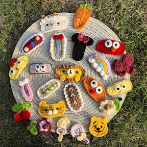 Könnte beinhalten: Eine Sammlung farbenfroher, handgehäkelter Haarspangen in verschiedenen Formen und Designs. Die Spangen umfassen eine Karotte, einen Bären, einen Regenbogen und andere skurrile Charaktere. Die Spangen sind auf einer runden, gewebten Matte vor grünem Gras angeordnet.