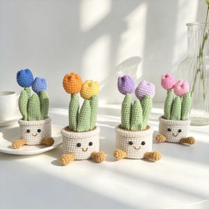 May include: Four crocheted potted plants, each featuring two colourful tulip flowers and green leaves. The white pots have embroidered smiling faces and small tan feet. The tulips are blue, orange, yellow, purple, and pink, crafted with yarn.