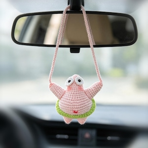 May include: A light pink crocheted starfish ornament with large white eyes and a green hula hoop around its waist. The figure is suspended by a long pink crocheted cord, designed to hang from a vehicle rearview mirror, featuring a soft texture.