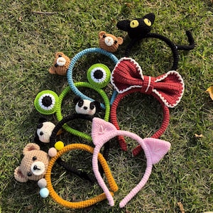 Pode incluir: Uma coleção de tiaras de crochê feitas à mão em vários designs, incluindo um gato preto, um laço vermelho, uma tiara de orelhas de gato rosa e um ursinho de pelúcia marrom. As tiaras estão sobre grama verde.
