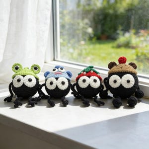 Puede incluir: Cuatro criaturas hechas a mano con ganchillo, con grandes ojos blancos y cuerpos negros. Cada una tiene un sombrero diferente: una rana verde, un sombrero azul con una cara, una fresa y un sombrero marrón con un pompón rojo. En un alféizar blanco.