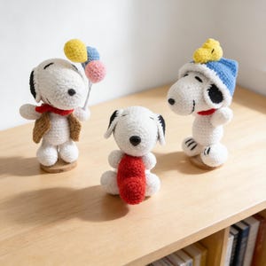 Könnte beinhalten: Drei handgefertigte Snoopy-Figuren aus Häkelarbeit. Einer hält Luftballons, ein anderer trägt einen blauen Hut und der dritte einen roten Schal. Alle sind weiß mit schwarzen Ohren und Nasen und stehen auf Holzsockeln.