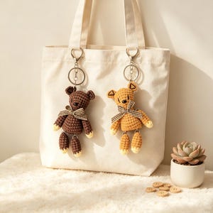 May include: A cream-colored canvas tote bag featuring two crocheted bear keychains attached to the straps. One bear is dark brown and the other is light tan, both wearing small checkered bows around their necks with silver metal hardware.