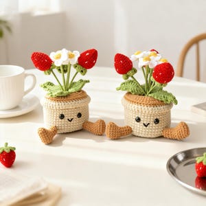 May include: Two crocheted strawberry plant pots with smiling faces and small legs. Each pot has red strawberries, white flowers, and green leaves. The pots are beige and brown, and sit on a white surface. A single strawberry is also visible.
