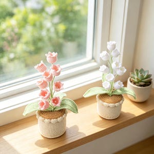Può includere: Due piante all'uncinetto in vaso con fiori a forma di campana in rosa tenue e bianco. Ogni pianta ha foglie verdi e poggia su un vaso testurizzato color crema con un bordo superiore beige. È visibile anche una piccola succulenta verde.