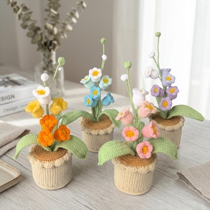 May include: Four crocheted potted flower arrangements featuring various colours including orange, yellow, blue, white, pink, and purple blossoms. Each plant has green stems and leaves, sitting in a beige crocheted pot with a brown top rim.