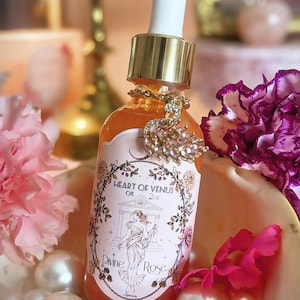 May include: A clear glass dropper bottle containing orange oil, labeled Heart of Venus Oil 2oz Divine Rose. The bottle features a gold cap and a white dropper, adorned with a gold-toned swan charm encrusted with small clear crystals around the neck.