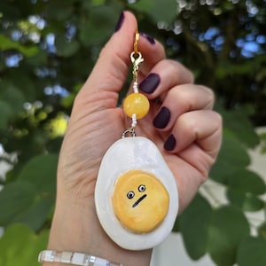 Könnte beinhalten: Ein handgefertigter, eiförmiger Anhänger mit überraschtem Gesicht, befestigt an einem goldfarbenen Verschluss. Der Anhänger ist weiß mit einem gelben Eigelb und schwarzen Gesichtszügen. Eine gelbe Perle und weiße Perlen befinden sich über dem Ei.