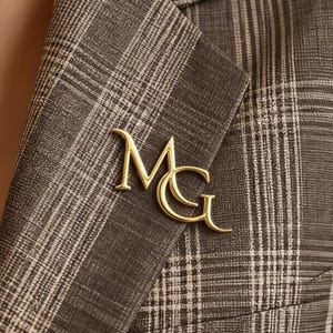 Op de afbeelding: Een goudkleurige metalen speld in de vorm van de letters "MG" is bevestigd aan de revers van een bruine geruite blazer. De blazer heeft een klassiek patroon in bruin- en beigetinten. De speld is een decoratief accessoire.
