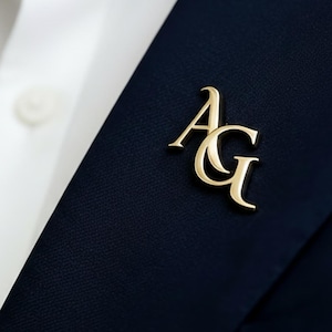 May include: A gold-colored lapel pin with the initials "AG" is affixed to a navy blue blazer. The pin is positioned on the lapel, near a white dress shirt. The image is a close-up shot, highlighting the pin's design and placement.