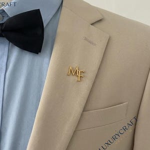 Puede incluir: Un blazer color beige con pajarita negra, camisa azul claro y un pin de solapa dorado con las iniciales "MF". El blazer tiene un bolsillo y está marcado con "LUXURYCRAFT".