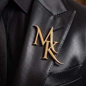 May include: A gold-toned metal brooch featuring the interlocking letters M and K in a stylized serif font, pinned to the lapel of a black blazer worn over a black shirt. The letters have a polished finish and sharp, pointed edges.
