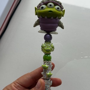 May include: A pen featuring a green alien character with three eyes, a purple outfit, and large black eyes. The pen's barrel is covered in rhinestones and adorned with small green alien figures. The pen is held in a hand.