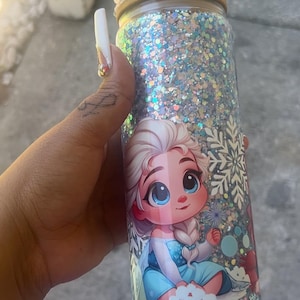 May include: A glitter-covered tumbler featuring a cartoon image of Elsa from Frozen. The tumbler has a wooden lid with Elsa and Olaf figurines on top. The tumbler is covered in silver and blue glitter.