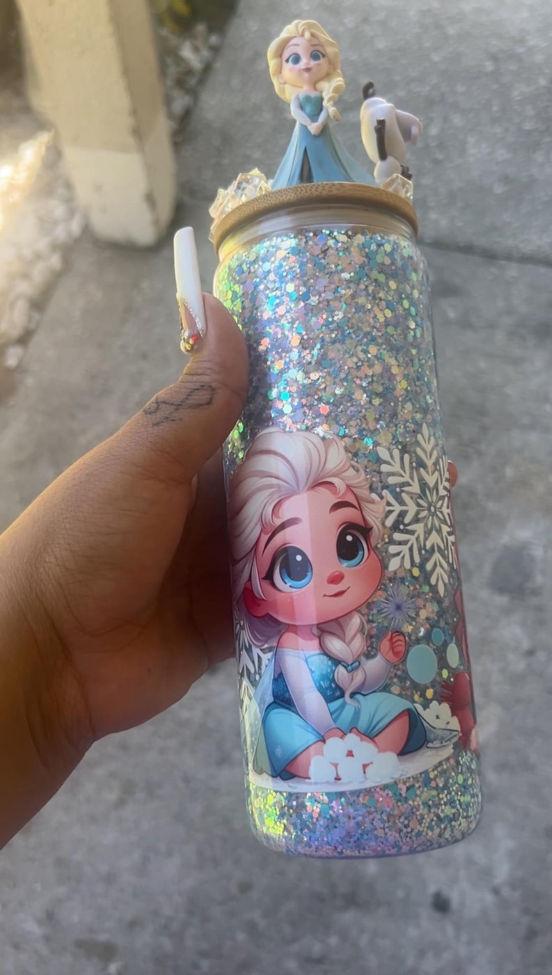 May include: A glitter-covered tumbler featuring a cartoon image of Elsa from Frozen. The tumbler has a wooden lid with Elsa and Olaf figurines on top. The tumbler is covered in silver and blue glitter.