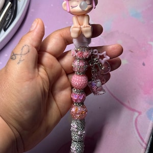 May include: A decorative pen with a pink figurine on top, featuring a bubble-blowing character. The pen's shaft is adorned with pink and silver beads and charms, creating a whimsical and stylish writing instrument.