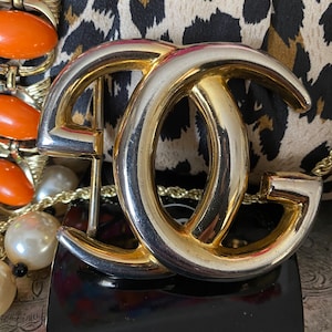K&ouml;nnte beinhalten: Eine goldfarbene G&uuml;rtelschnalle aus Metall mit einem doppelten 'C'-Design. Die Schnalle befindet sich auf einem Leopardenmuster-Hintergrund.