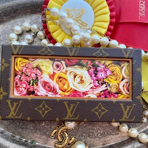 May include: A brown leather frame with a floral print inside. The frame is decorated with a gold LV logo. The frame is surrounded by pearls and a yellow ribbon.