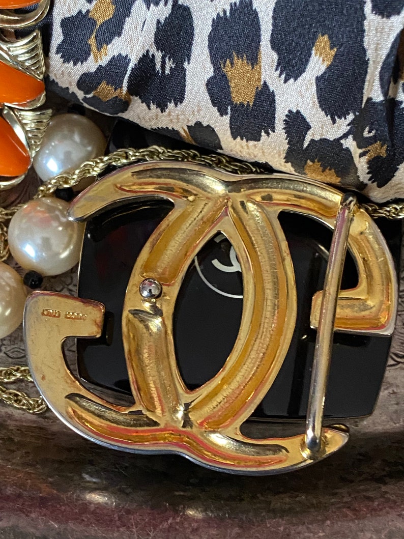 K&ouml;nnte beinhalten: Eine goldfarbene G&uuml;rtelschnalle aus Metall mit einem Doppel-'C'-Design. Die Schnalle hat eine polierte Oberfl&auml;che und einen silberfarbenen Rand.