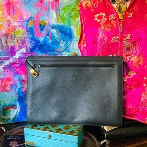 May include: A black leather clutch with a gold zipper and logo. The clutch is rectangular and has a textured surface. A turquoise box with gold accents and a vintage photo are in the foreground. A pink floral vest is in the background.