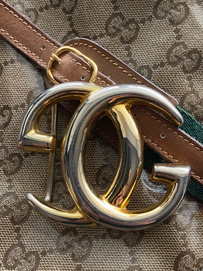 K&ouml;nnte beinhalten: Eine goldene und silberne, ineinandergreifende G-Schnalle mit einem braunen Lederg&uuml;rtel. Die Schnalle ist ein beliebtes Designelement f&uuml;r Luxusmodeaccessoires.