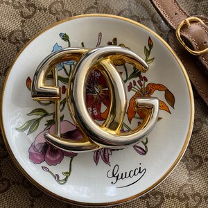 K&ouml;nnte beinhalten: Ein goldfarbenes Doppel-'G'-Logo auf einer wei&szlig;en Porzellanschale mit Blumenmuster und dem Wort "Gucci" in kursiver Schrift.