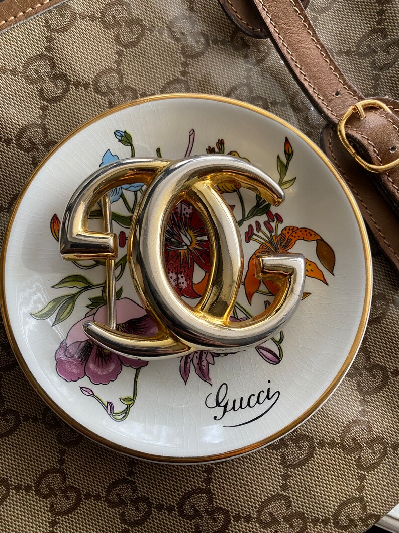 K&ouml;nnte beinhalten: Ein goldfarbenes Doppel-'G'-Logo auf einer wei&szlig;en Porzellanschale mit Blumenmuster und dem Wort "Gucci" in kursiver Schrift.