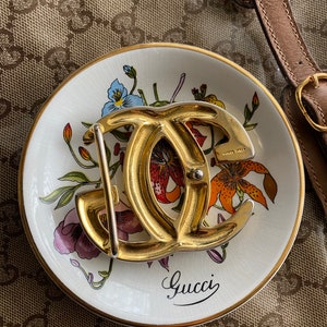 K&ouml;nnte beinhalten: Eine goldfarbene Gucci-G&uuml;rtelschnalle mit einem Doppel-G-Logo. Die Schnalle befindet sich auf einem wei&szlig;en Teller mit einem Blumenmuster und dem Wort "Gucci" in schwarzer Schrift.