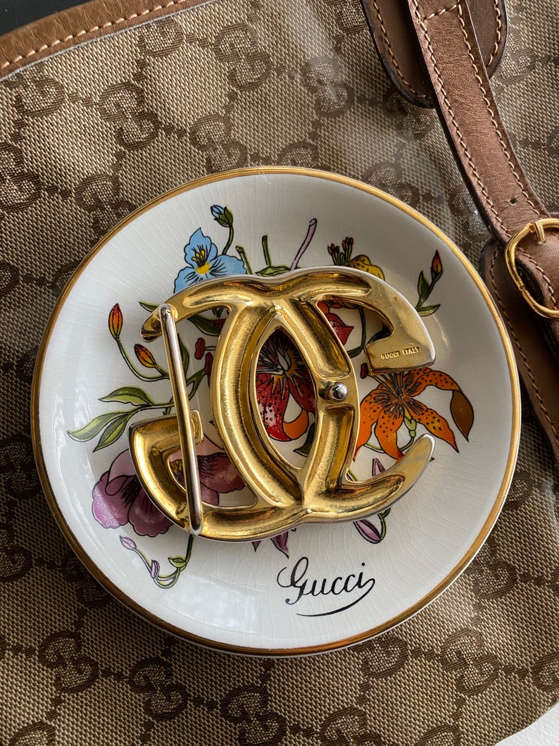K&ouml;nnte beinhalten: Eine goldfarbene Gucci-G&uuml;rtelschnalle mit einem Doppel-G-Logo. Die Schnalle befindet sich auf einem wei&szlig;en Teller mit einem Blumenmuster und dem Wort "Gucci" in schwarzer Schrift.