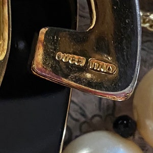 K&ouml;nnte beinhalten: Nahaufnahme eines goldfarbenen Metallanh&auml;ngers mit schwarzem Emaille-Design. Der Anh&auml;nger tr&auml;gt die Aufschrift "GUCCI ITALY" auf der R&uuml;ckseite.