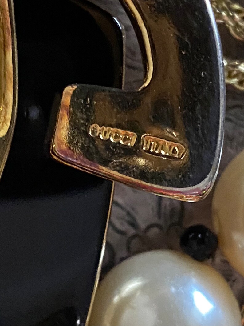 K&ouml;nnte beinhalten: Nahaufnahme eines goldfarbenen Metallanh&auml;ngers mit schwarzem Emaille-Design. Der Anh&auml;nger tr&auml;gt die Aufschrift "GUCCI ITALY" auf der R&uuml;ckseite.
