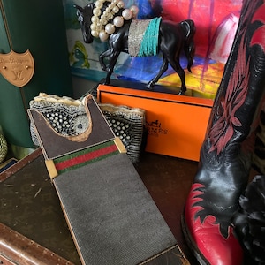 May include: A black horse figurine adorned with pearl necklaces and turquoise and silver beaded accents. A vintage Gucci case with green and red stripes, a pair of black and red cowboy boots, and an orange box are also visible.