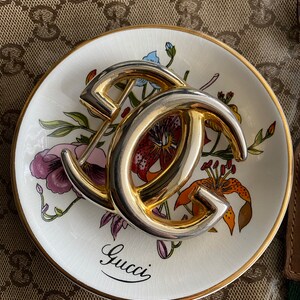 K&ouml;nnte beinhalten: Ein goldfarbenes Metall-Gucci-Logo auf einer wei&szlig;en Porzellanschale mit Blumenmuster. Die Schale hat einen goldenen Rand.