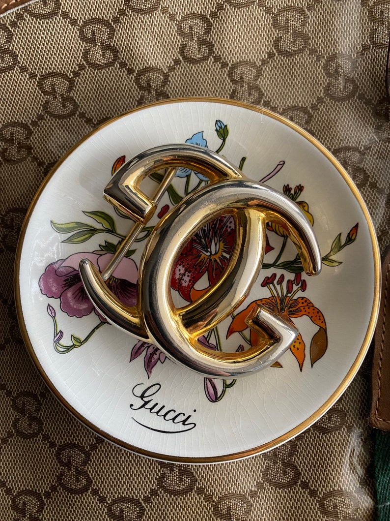 K&ouml;nnte beinhalten: Ein goldfarbenes Metall-Gucci-Logo auf einer wei&szlig;en Porzellanschale mit Blumenmuster. Die Schale hat einen goldenen Rand.