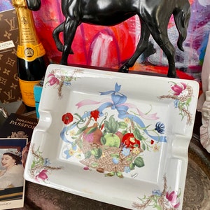May include: A rectangular white ashtray with a fruit basket design, including apples, pears, and pomegranates, decorated with blue ribbons and floral patterns. The ashtray has a decorative border and the word "Gucci" is printed on the side.