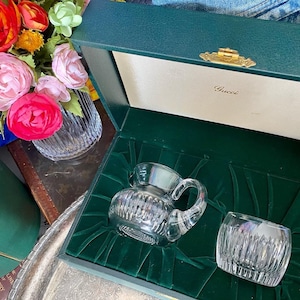 May include: A crystal creamer and tumbler set inside a dark green box with the Gucci logo. The creamer has a handle and a spout. The box is lined with green velvet. A vase of colorful flowers is in the background.