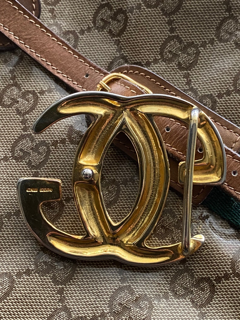 K&ouml;nnte beinhalten: Eine goldfarbene G&uuml;rtelschnalle aus Metall mit einem Doppel-'G'-Design. Die Schnalle ist an einem braunen Lederg&uuml;rtel mit einem gr&uuml;n-braunen gemusterten Stoff befestigt.
