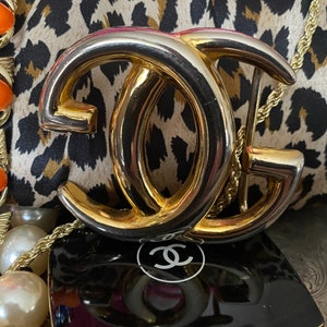 K&ouml;nnte beinhalten: Eine goldfarbene Doppel-'C'-Schnalle mit silberfarbenem Finish. Die Schnalle liegt auf einem Leopardenmusterstoff mit einem schwarzen und wei&szlig;en Chanel-Logo im Hintergrund.