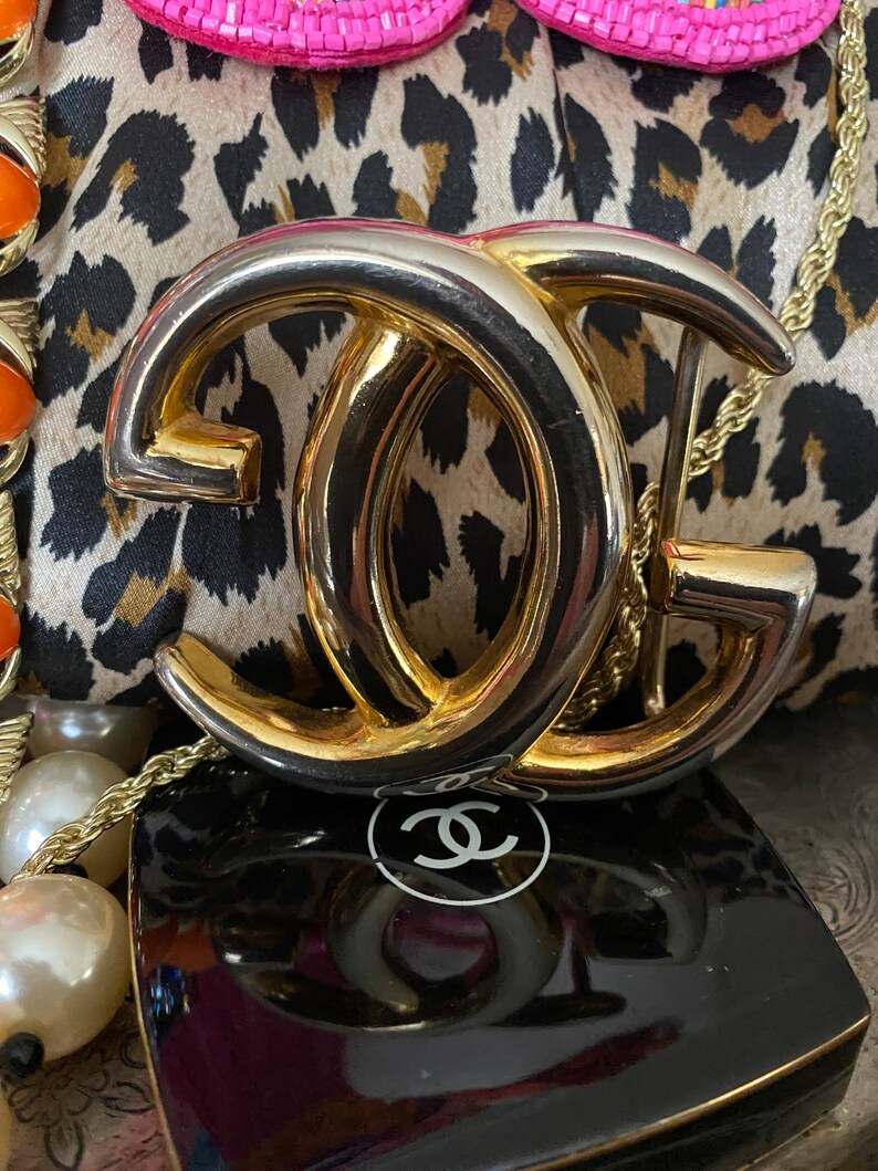 K&ouml;nnte beinhalten: Eine goldfarbene Doppel-'C'-Schnalle mit silberfarbenem Finish. Die Schnalle liegt auf einem Leopardenmusterstoff mit einem schwarzen und wei&szlig;en Chanel-Logo im Hintergrund.