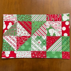 May include: A rectangular Christmas-themed quilted placemat. The patchwork design features red, green, and white squares and triangles with holiday patterns like snowmen, candy canes, and polka dots. The edges are neatly stitched.