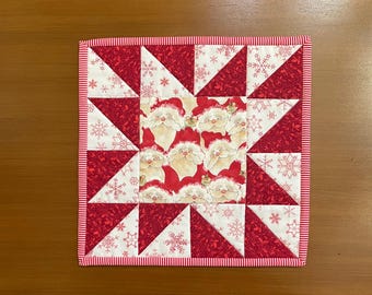 May include: Square Christmas-themed quilted placemat. The center features a pattern of Santa Claus faces. The surrounding triangles alternate between red snowflake and floral patterns. A red and white striped border frames the design.