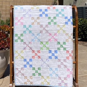 May include: A white quilt with a colorful patchwork design, displayed on a wooden rack. The quilt features a repeating pattern of squares in pastel colors like blue, pink, green, and yellow. The edges are finished with a light blue border.