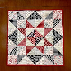 May include: A square quilt with a patchwork design featuring triangles and squares in red, black, white, and gray fabrics. The quilt has a red border and a variety of patterns, including hearts, polka dots, and script.