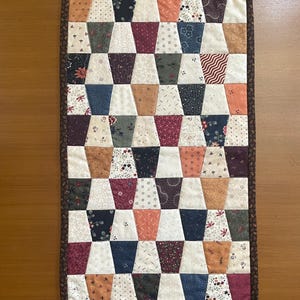 May include: A rectangular quilted table runner featuring a repeating pattern of trapezoidal fabric patches in various colors including cream, navy blue, dark red, olive green, and burnt orange, all bordered by a dark brown fabric with a floral print.